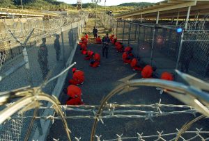 Detainees in orange jumpsuits sit in a holding area at Camp X-Ray of Naval Base Guantanamo Bay (January 11, 2002). Image via Reuters File.